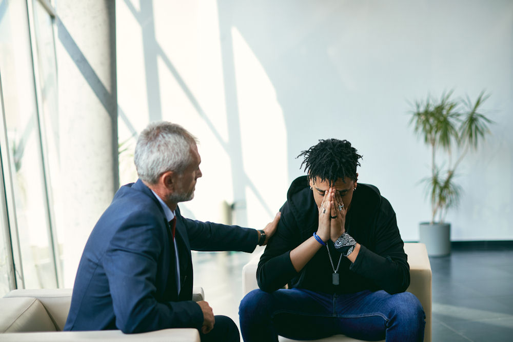 older man sitting beside a younger guy in distress and offering him support