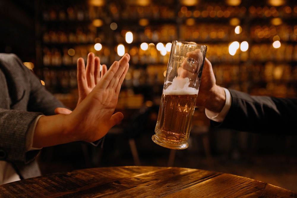 person refusing a beer by placing their hands in front of it while another person hands a glass of beer over the table