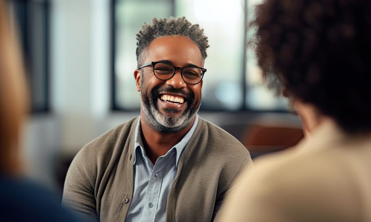 Group therapy and support. The focus is on a middle aged African American man in eyeglasses. A group of people around support him. He is happy.